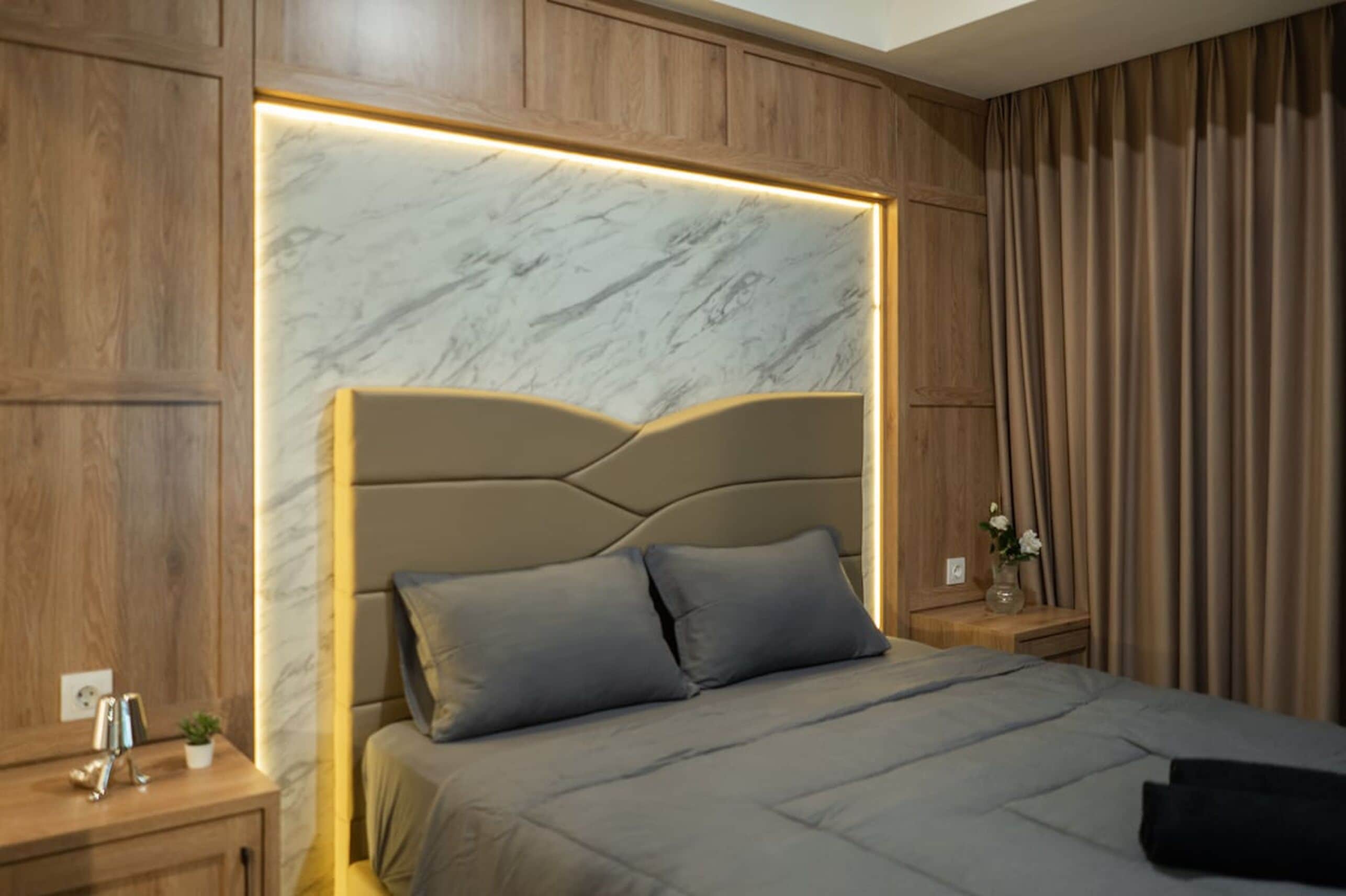 Un dormitorio moderno con cabecera de mármol, paneles de madera, una cama con ropa de cama gris, una mesita de noche con un jarrón de flores y una cortina en la ventana.