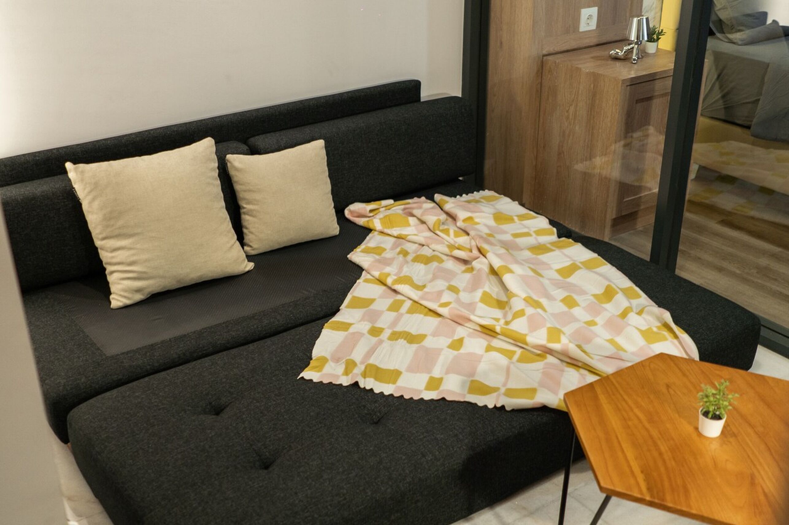 Una sala de estar moderna con un sofá gris oscuro, dos cojines color beige y una manta a cuadros. Hay una pequeña mesita de madera con una planta en maceta.
