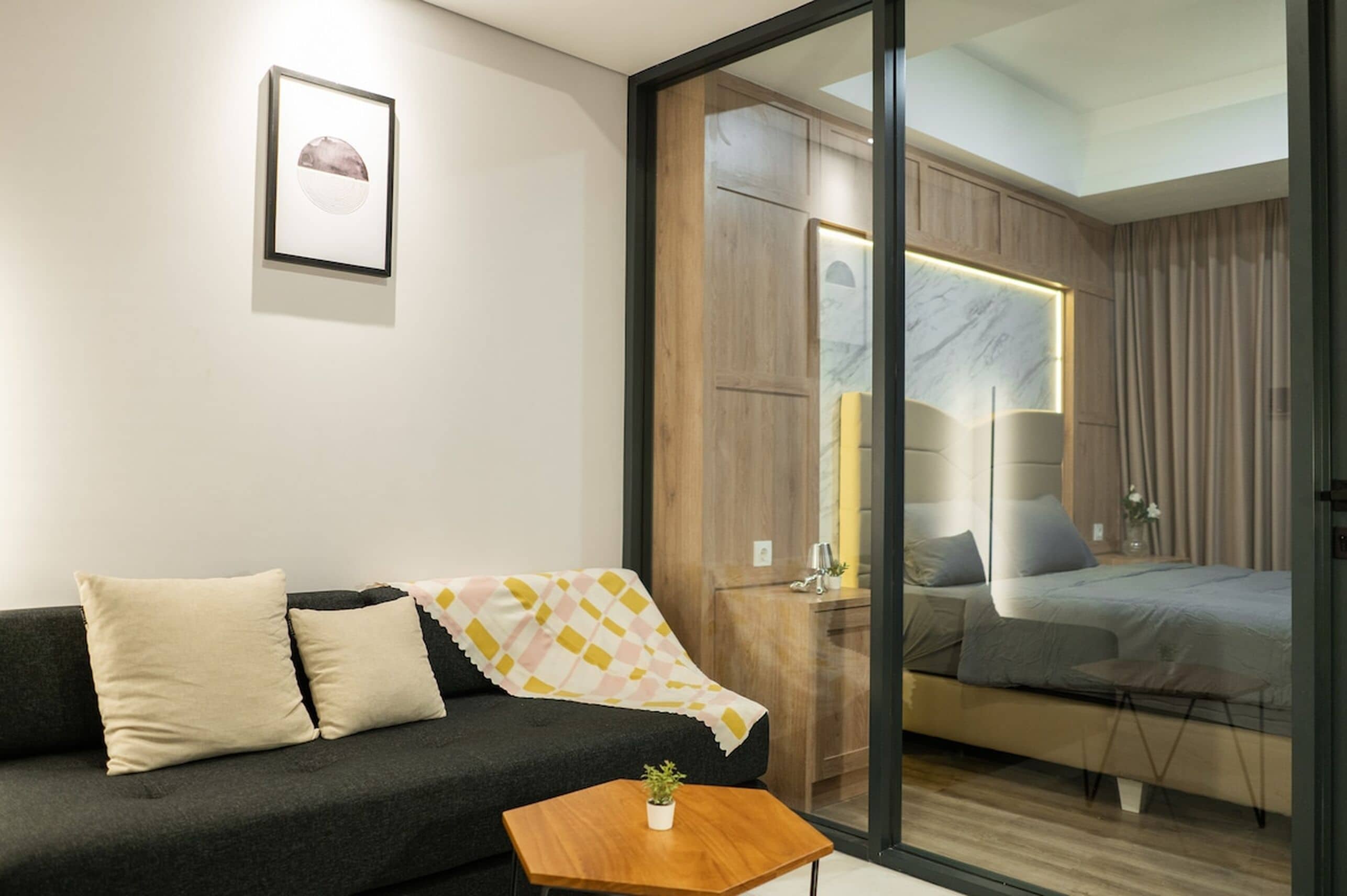 A modern room with a sofa, a small table with a plant, a bed with a headboard, and a sliding door leading to another room.