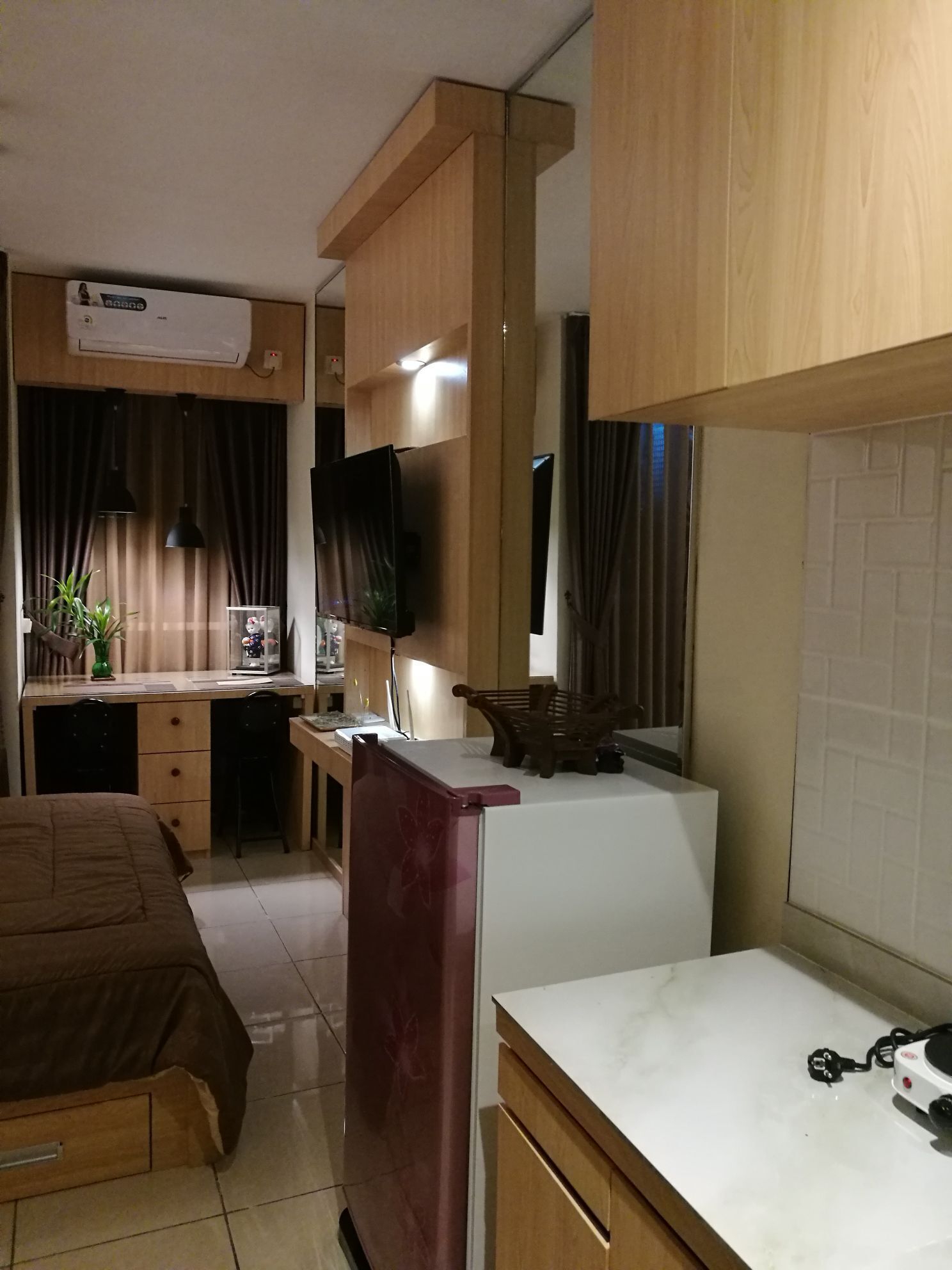 A compact hotel room with a bed, a desk with a computer, a small kitchenette, and a TV.