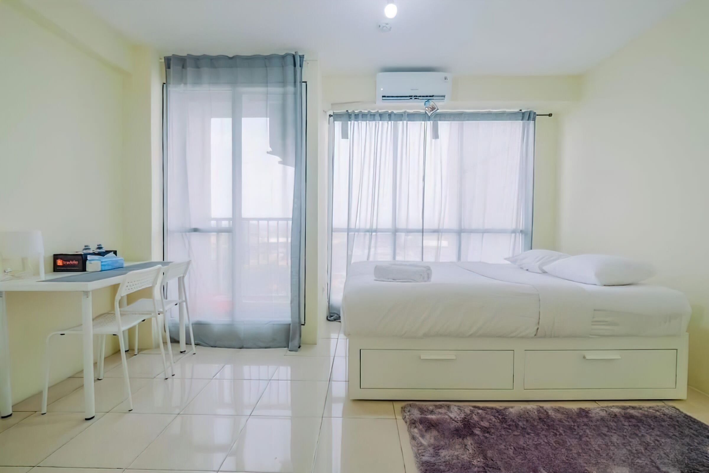 A hotel room with a bed, a desk with chairs, a window with curtains, and an air conditioning unit.