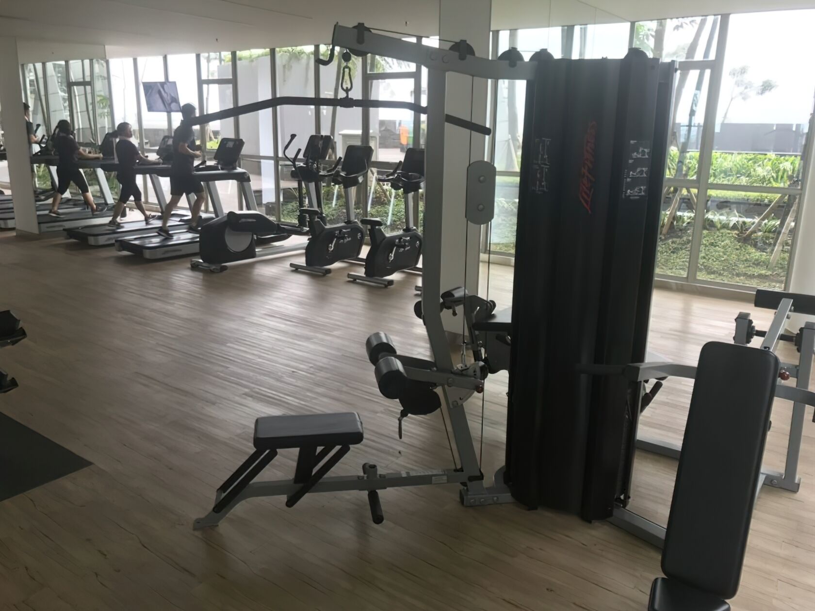 A gym with various exercise equipment, large windows, and a wooden floor.