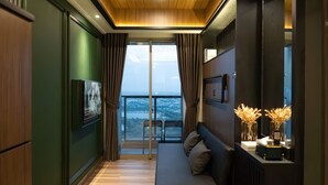 A modern room with a wooden floor, a sofa with cushions, a TV, and a view of a balcony with chairs. - Seaview GoldCoast Residence in Jakarta (Jakarta)