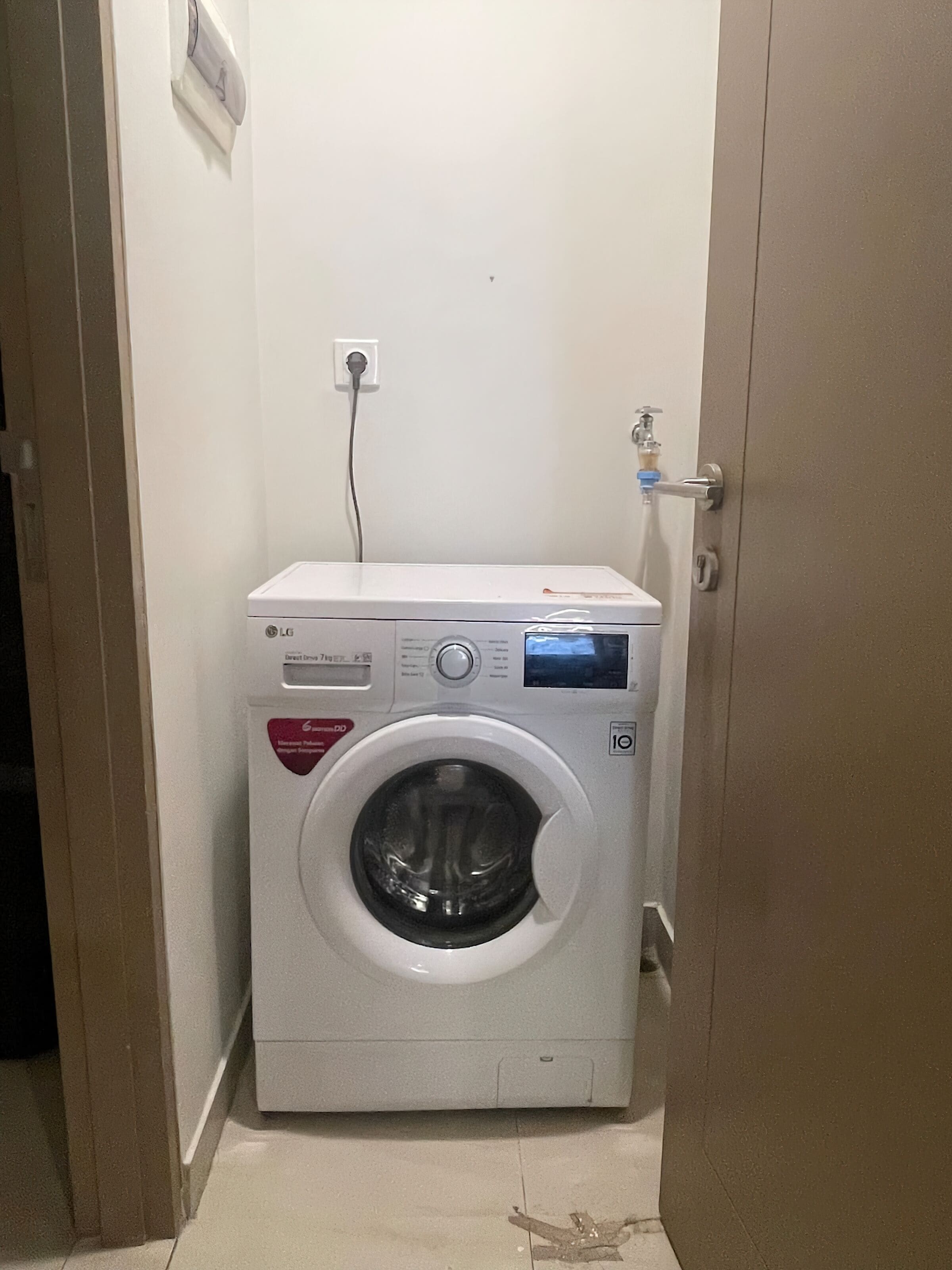 A compact, white front-loading washing machine in a small, enclosed laundry room with a door on one side and a power outlet on the wall.