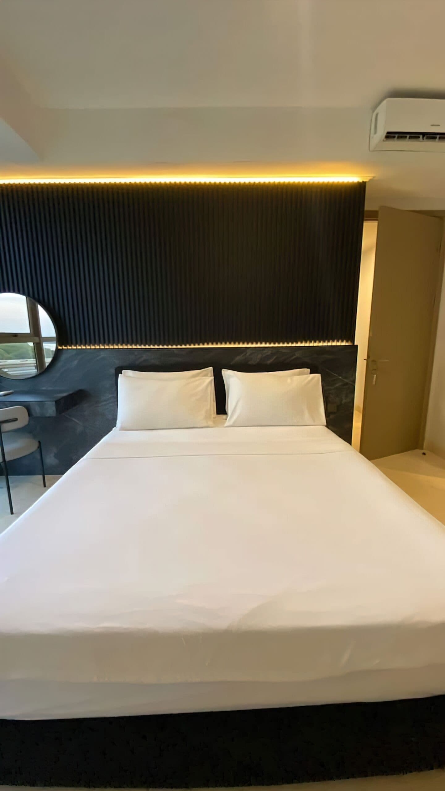 A modern hotel room with a large bed, a headboard with vertical slats, an air conditioning unit above the bed, and a small desk with a chair.