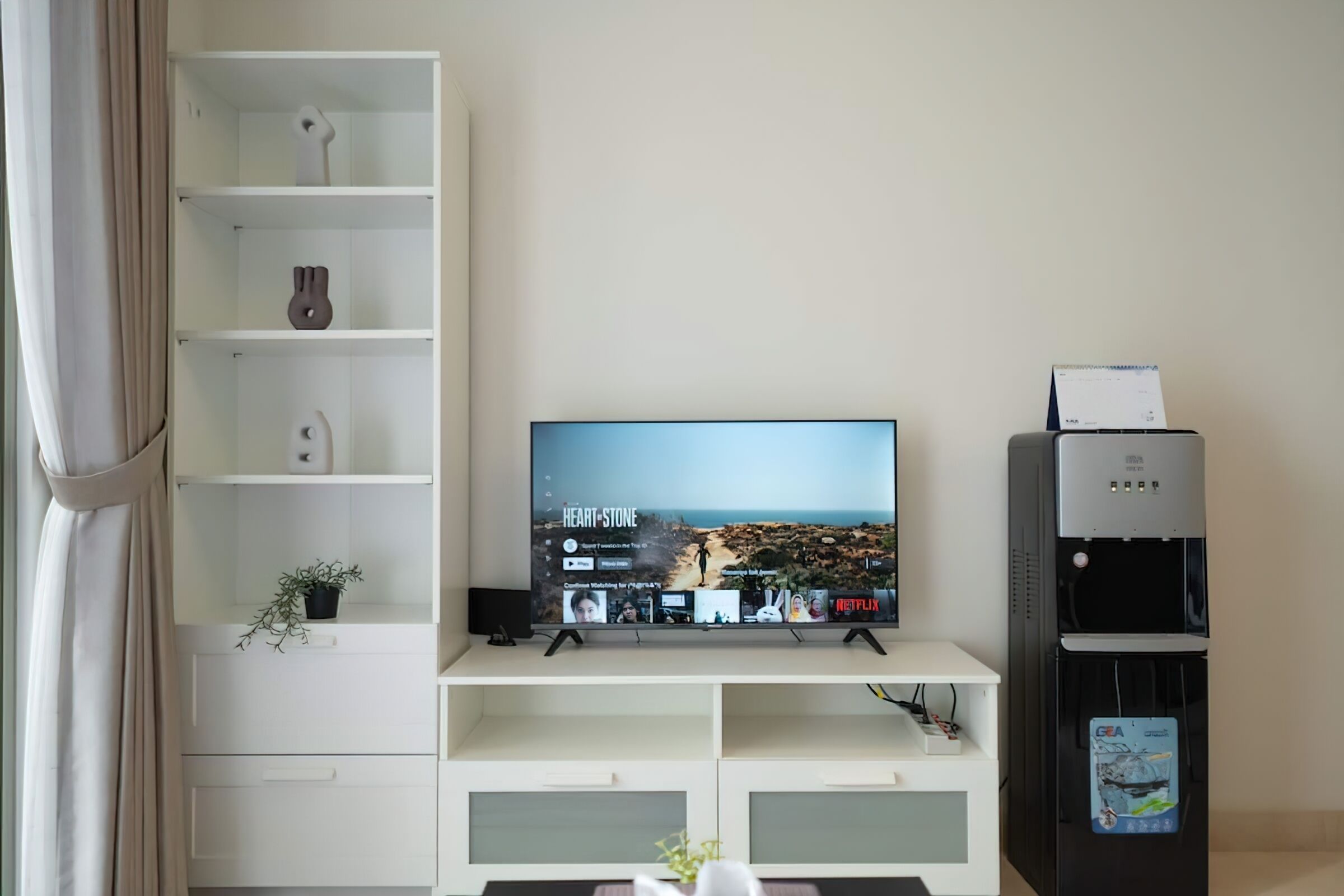 Een woonkamer met een televisie op een standaard, een waterdispenser en een plank met decoratieve objecten.
