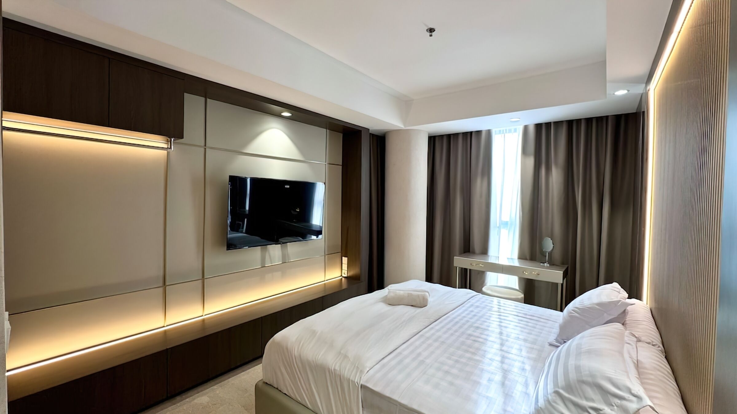 A modern hotel room with a large bed, a flat-screen TV mounted on the wall, a vanity with a mirror, and a window with curtains.