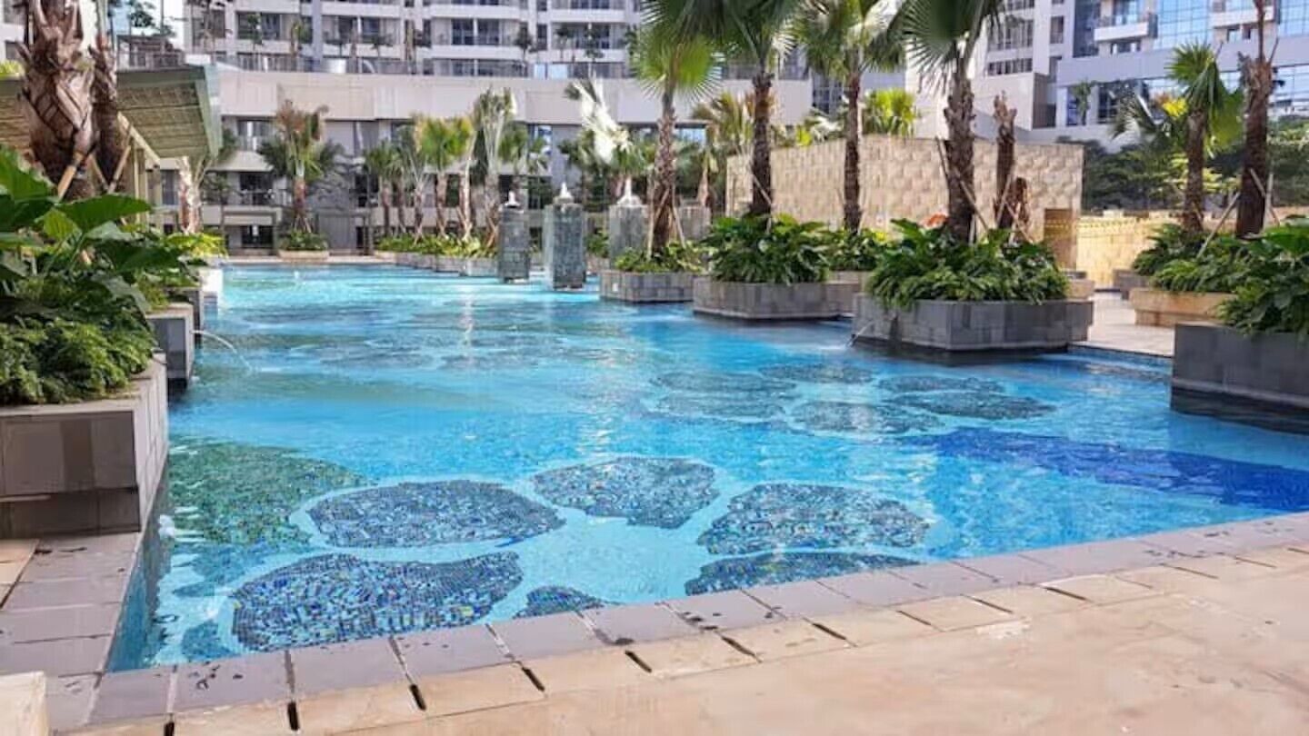 Ein Schwimmbecken mit Mosaikgestaltung, umgeben von Palmen und Pflanzgefäßen, mit modernen Gebäuden im Hintergrund.