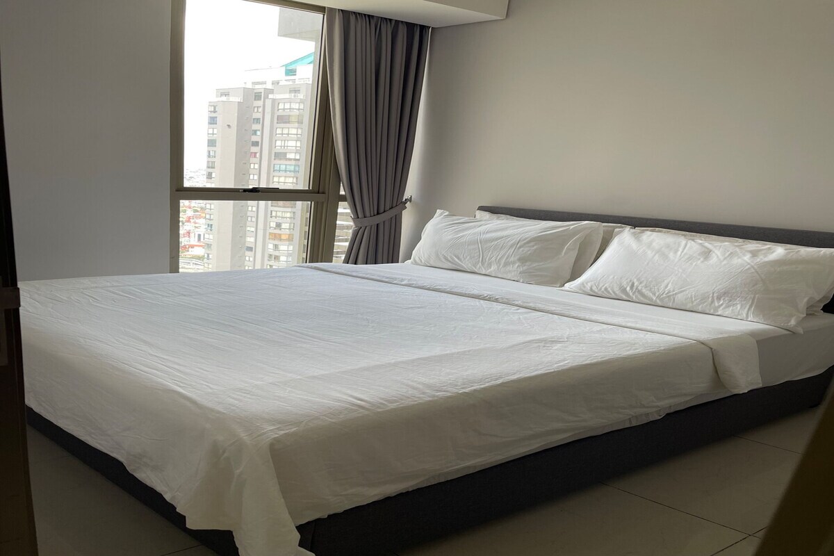 A hotel room with a large bed, a window with curtains, and a view of high-rise buildings.