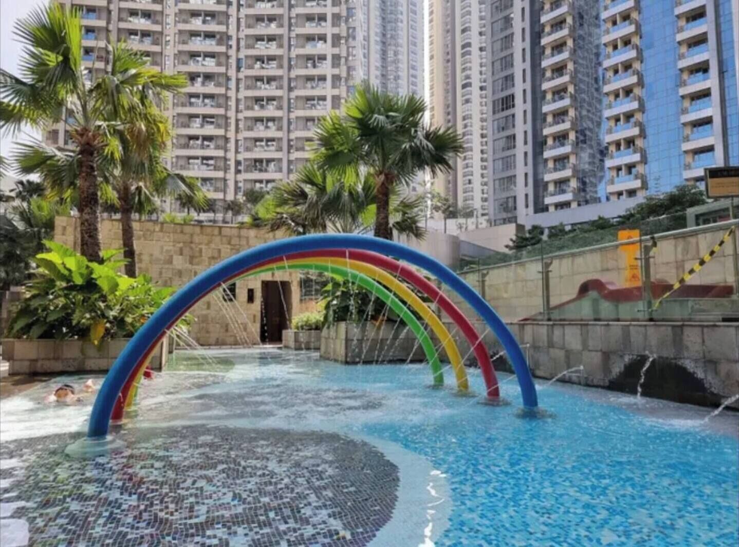 A swimming pool with a colorful arch structure, surrounded by tall residential buildings and palm trees.