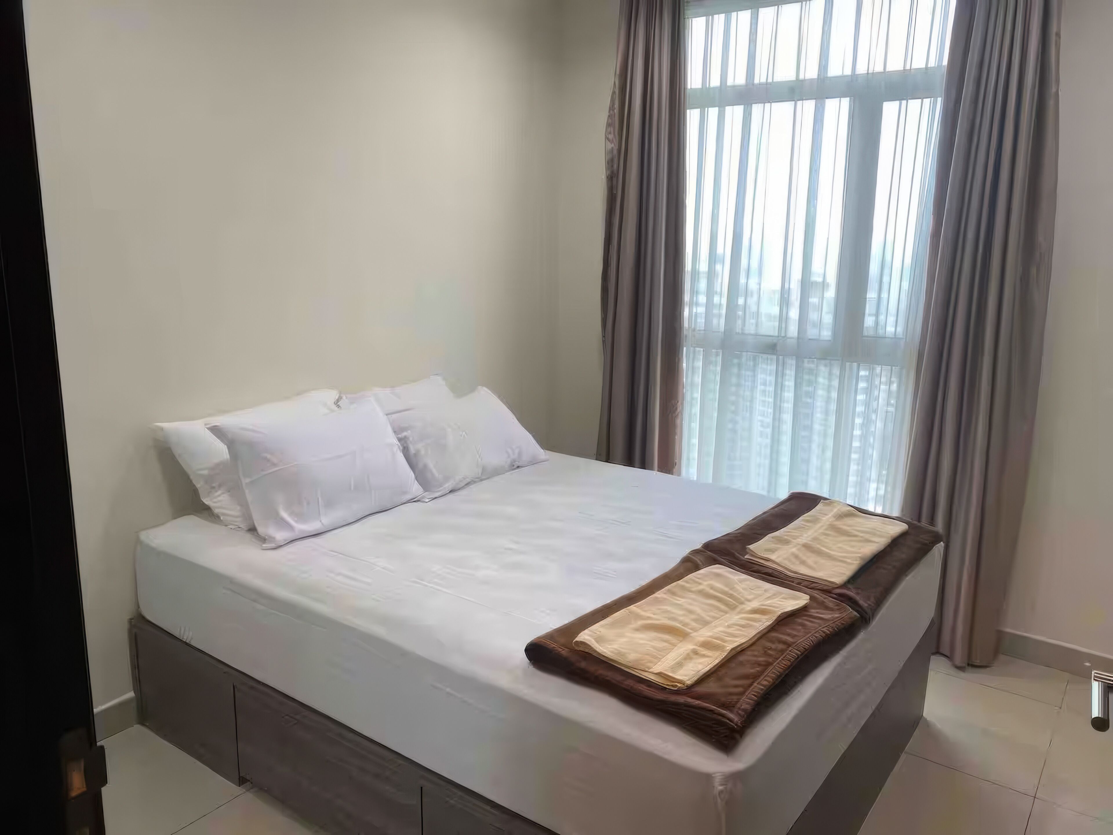 A neatly made bed with white linens and a brown throw blanket, a window with curtains, and a tiled floor.