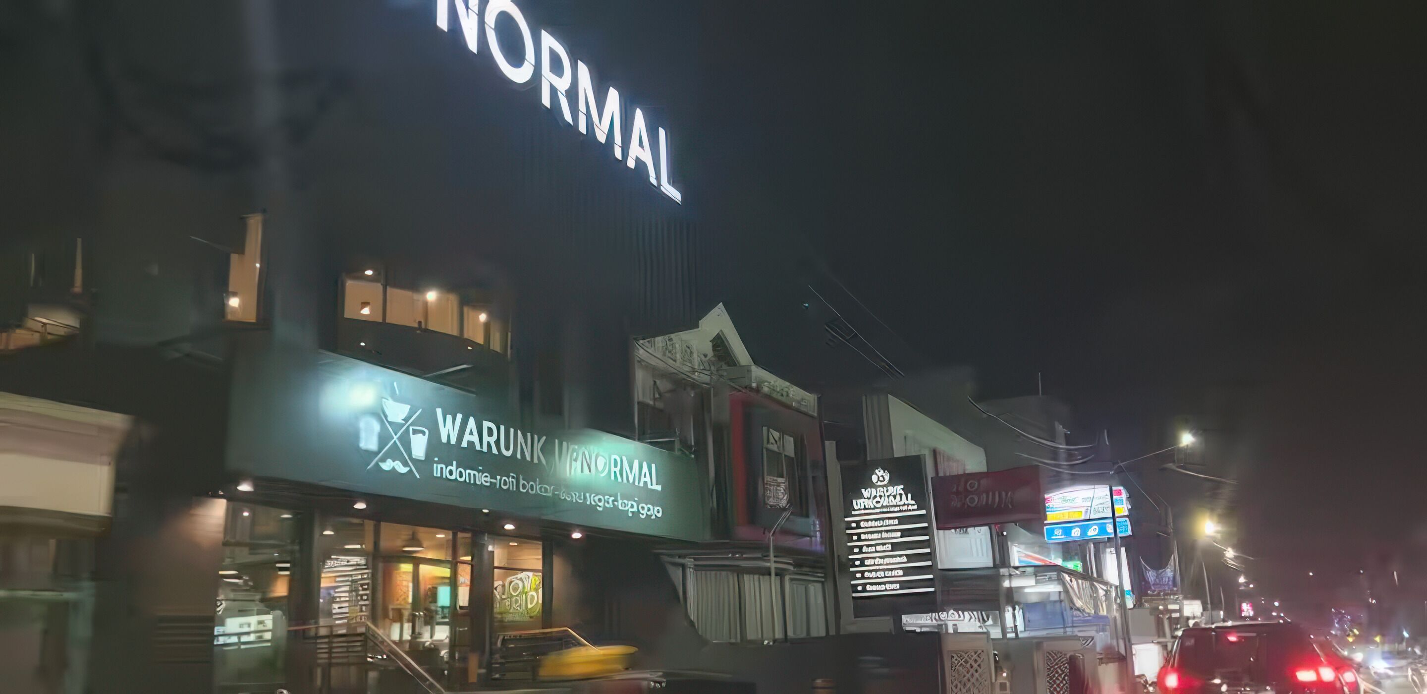 A building with a sign that reads "WARUNK WAROMAL" and another sign above it that says "NORMAL".