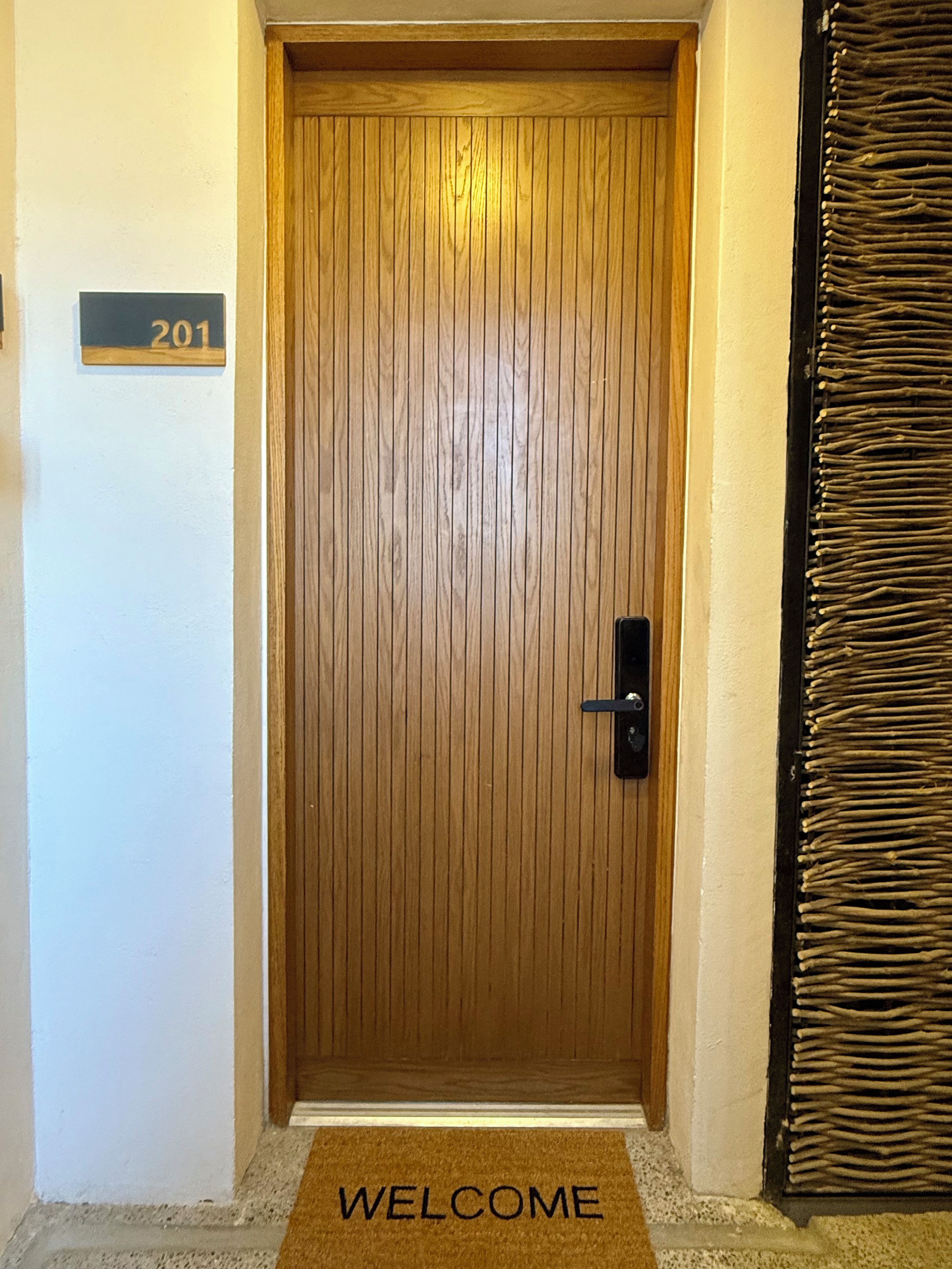 A wooden door with a 'WELCOME' mat at the entrance, a door number sign '201', and a textured wall to the right.