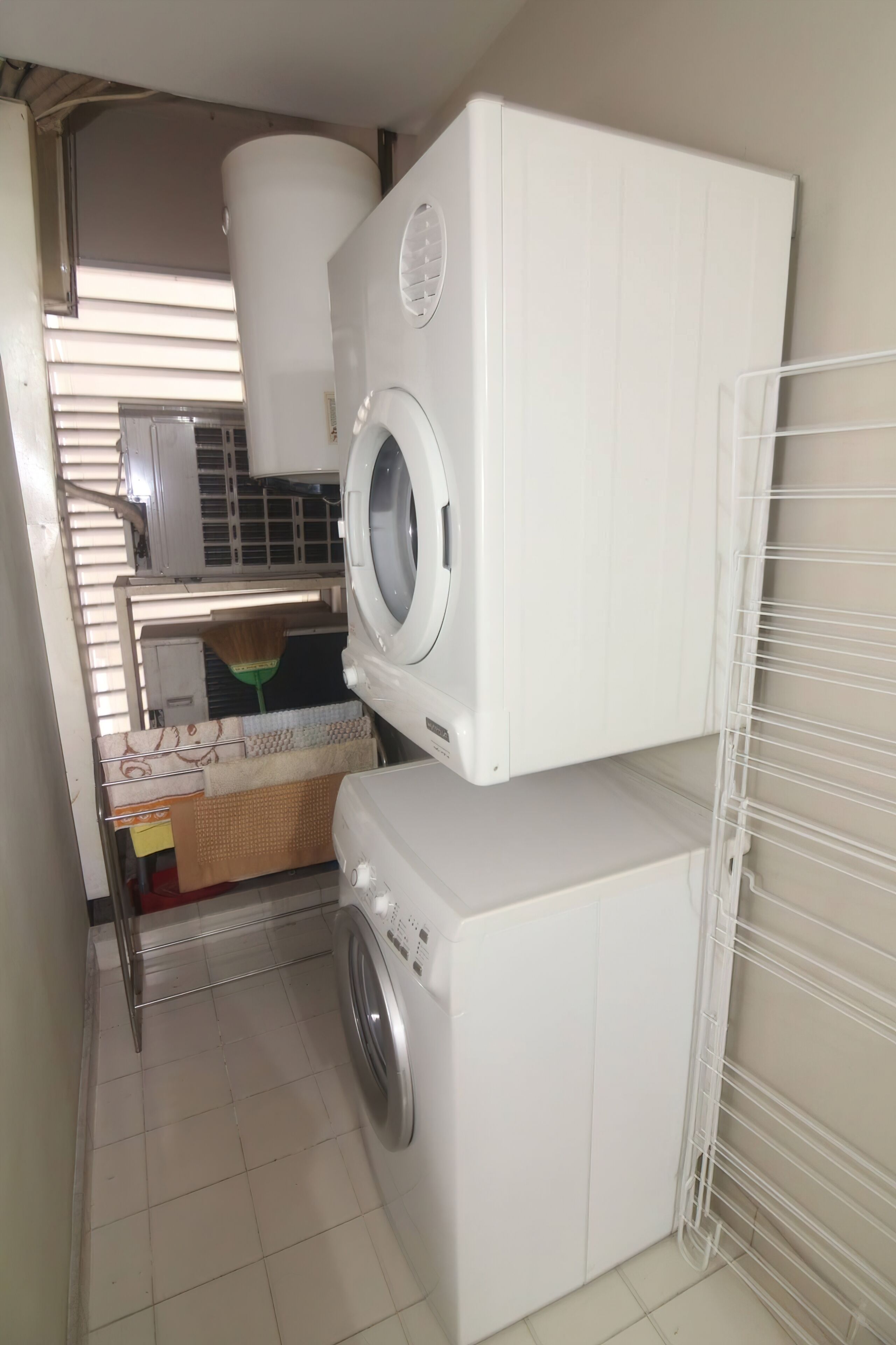 A compact laundry room with stacked appliances, a drying rack, and a broom.