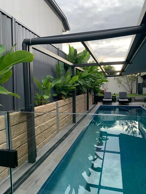A modern outdoor pool area with a glass enclosure, surrounded by plants and a stone wall. - Aurora Villa (Forest Lake)