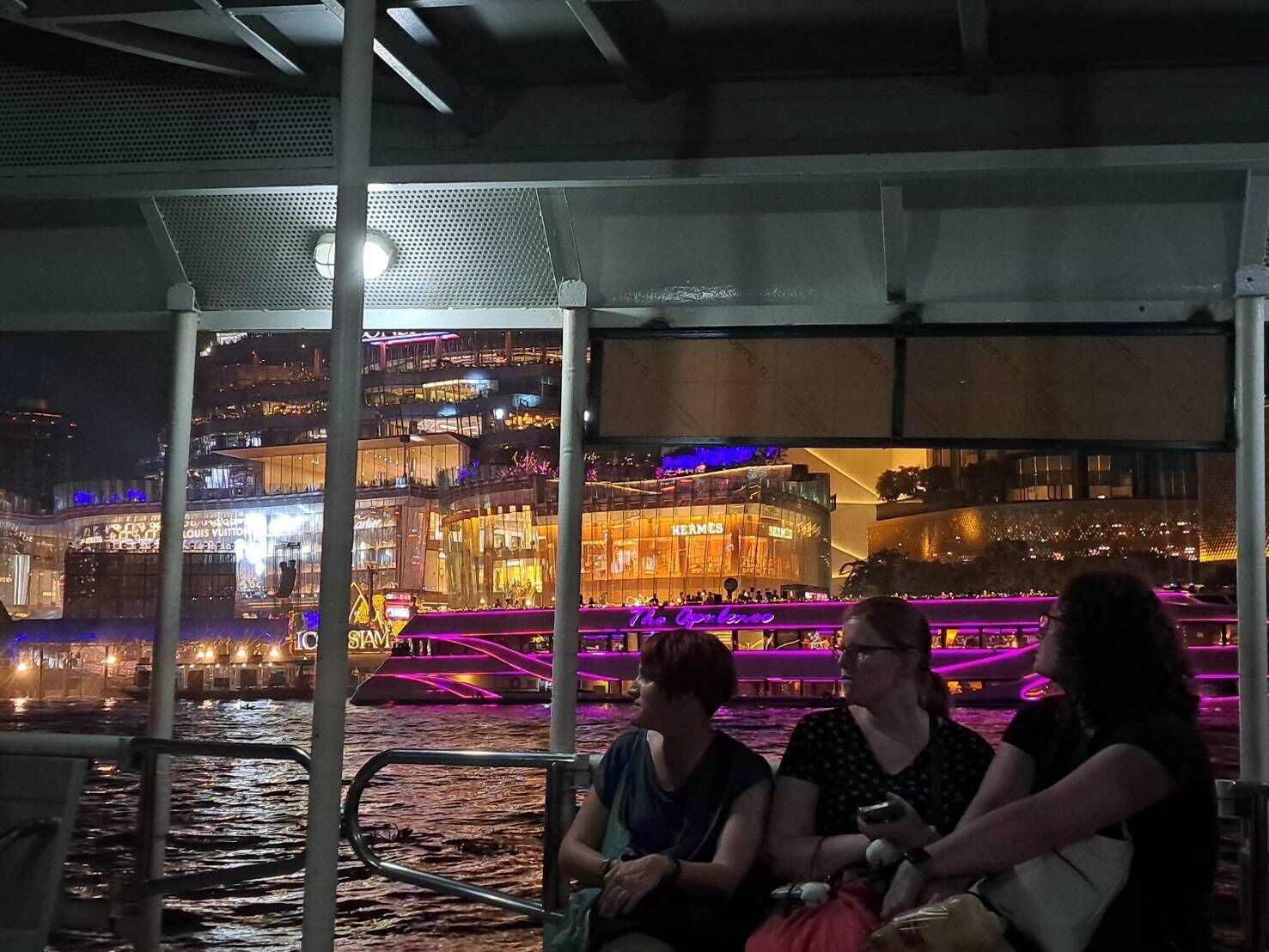 Vista noturna de uma área à beira-mar, com edifícios iluminados e um espaço coberto com assentos.