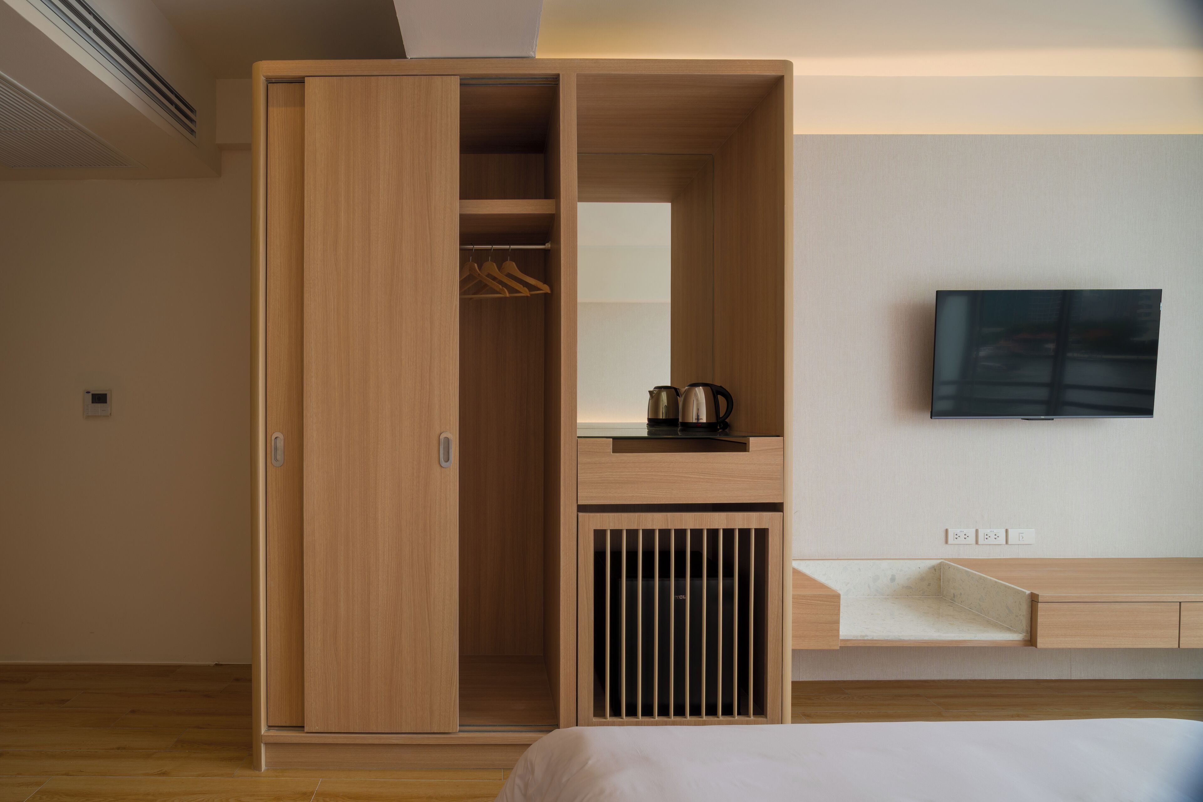 A modern hotel room with a wooden wardrobe, a flat-screen TV mounted on the wall, and a bedside table with a kettle.