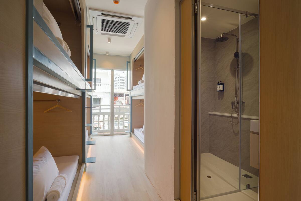 Ein Hotelzimmer mit Duschkabine aus Glas, einer Couch mit zusammengerolltem Handtuch und Blick auf die Stadt durch ein großes Fenster.