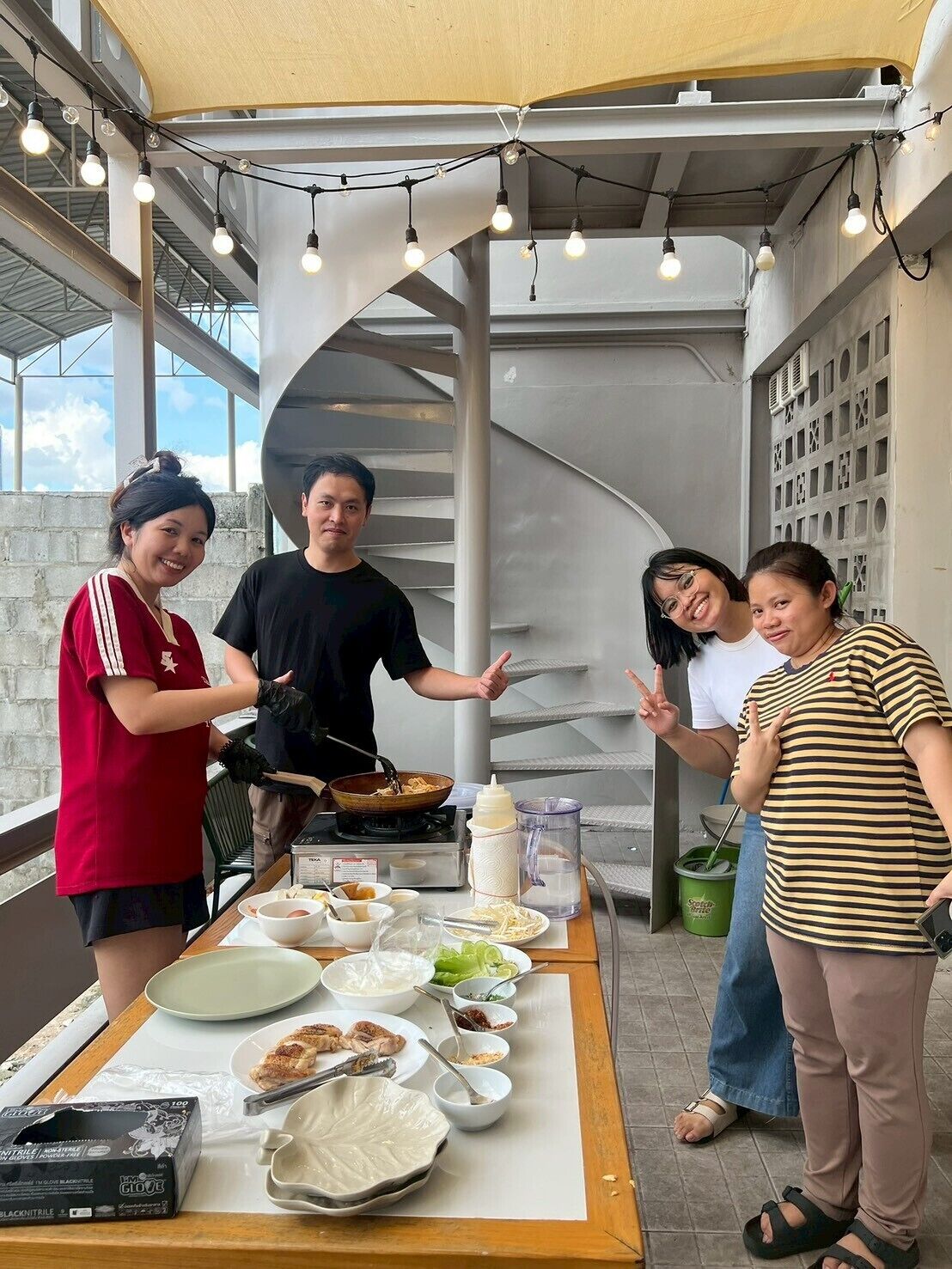 A group of people around a table with various dishes and cooking utensils, under a canopy with string lights.