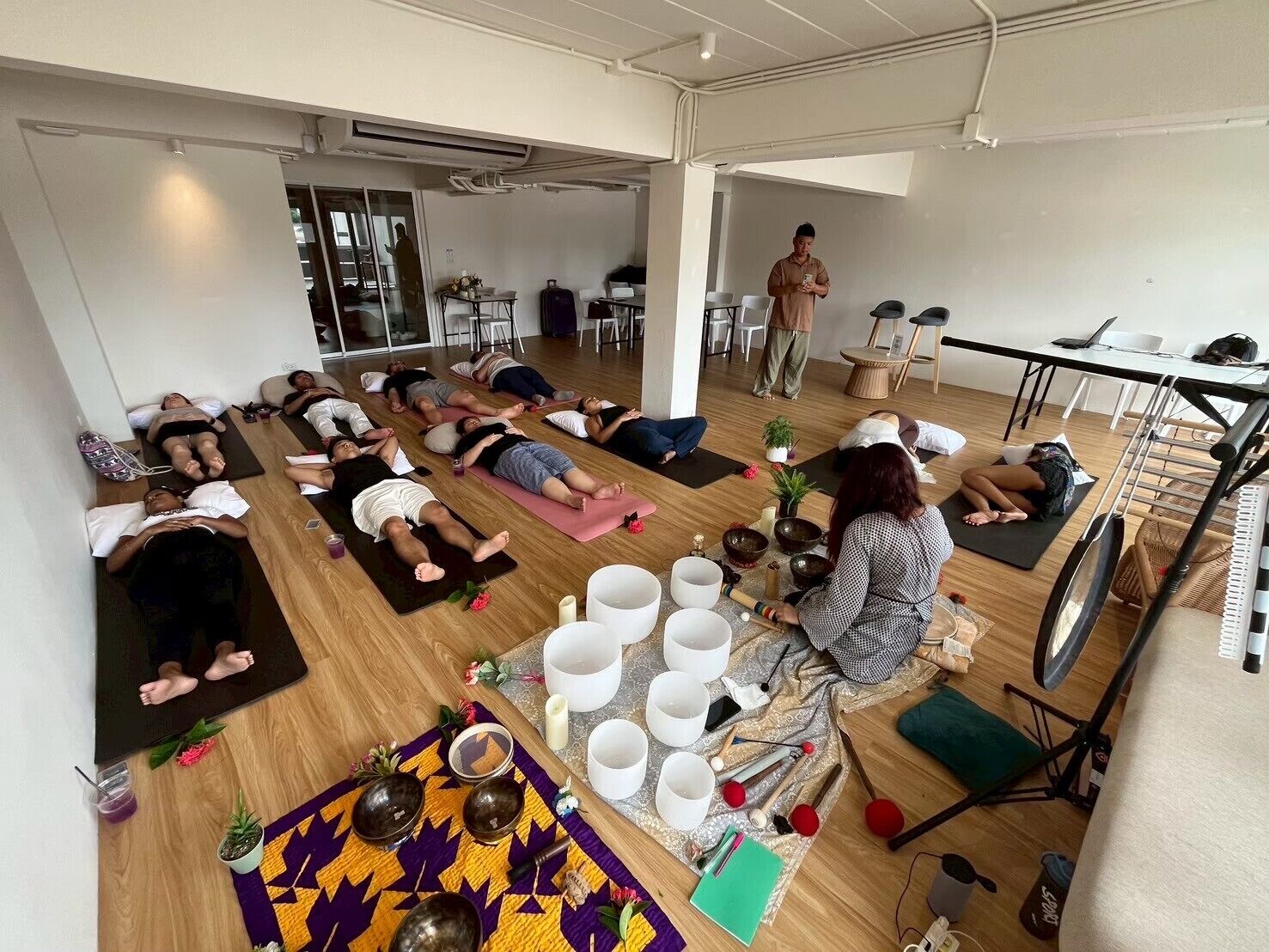 Clase de yoga en una habitación amplia con suelos de madera, una mesa central con cuencos y tazas, y participantes acostados sobre esterillas.