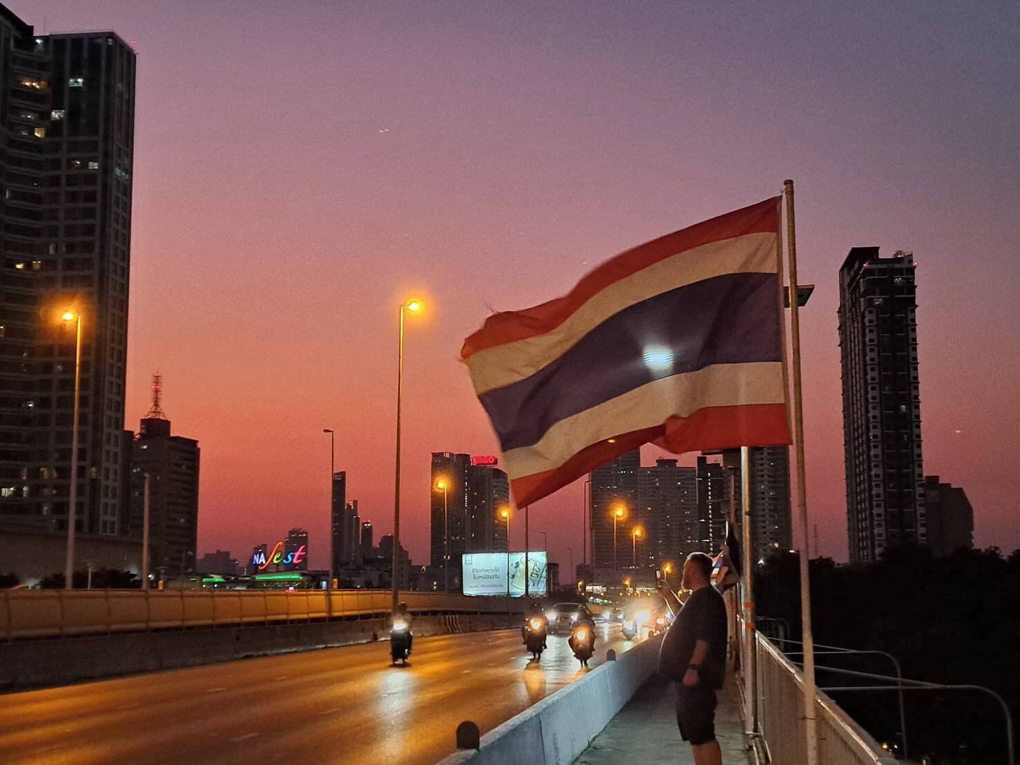 A cityscape at dusk with a prominent flag on a pole, tall buildings, and a busy road with vehicles.