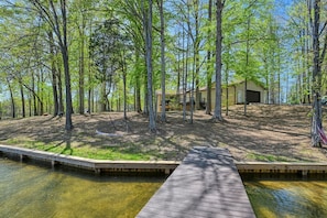 A cabin surrounded by tall trees, a dock extending into a clear body of water, and a small building nearby.