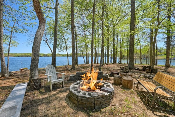 A campsite with a stone fire pit, wooden benches, and a chair by a lake.