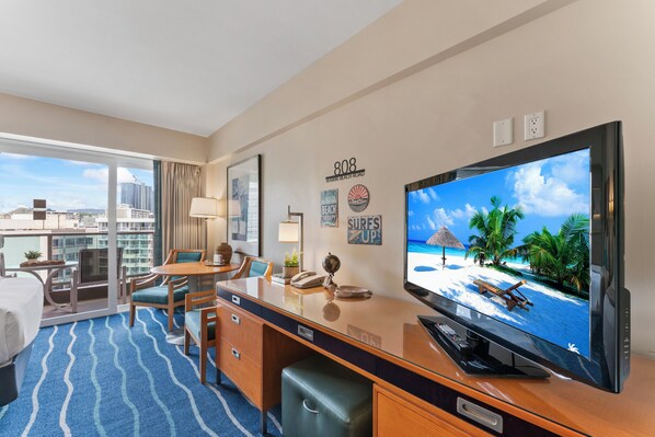 A hotel room with a large TV, a desk, a chair, a bed, and a view of the city.