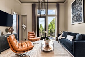 Un salon moderne avec des fauteuils en cuir, un canapé foncé, une table basse en verre et une grande fenêtre donnant sur la verdure.