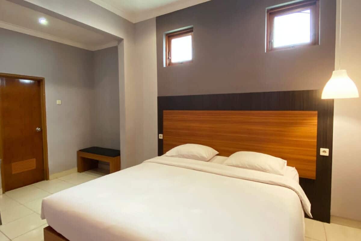 A bedroom with a large bed, two windows, a wooden headboard, a bedside table, and a door.