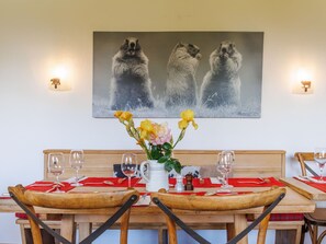 Une table à manger avec des chaises, un vase de fleurs et un tableau représentant trois marmottes accroché au mur.