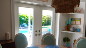 A dining area with a table set for four, a wicker chandelier, and a view of a pool and greenery outside.