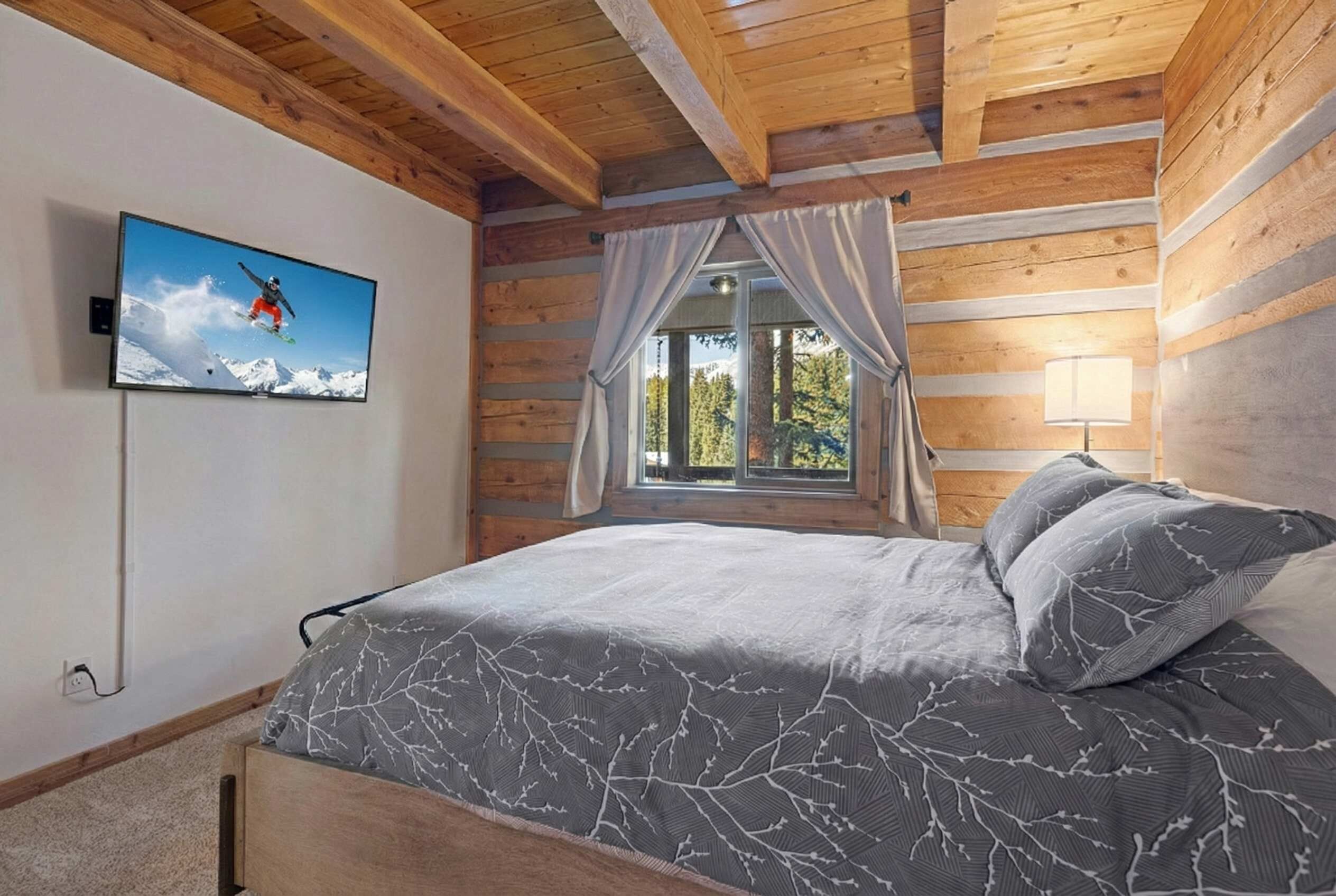 Une chambre à coucher avec un lit, un téléviseur fixé au mur, une fenêtre avec des rideaux et une lampe.