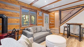 A cozy cabin interior with a fireplace, a sofa, a round ottoman, and a dining area with stools.