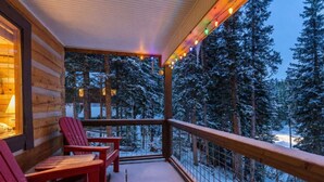 A wooden cabin with a balcony, red Adirondack chairs, and a snowy forest view.
