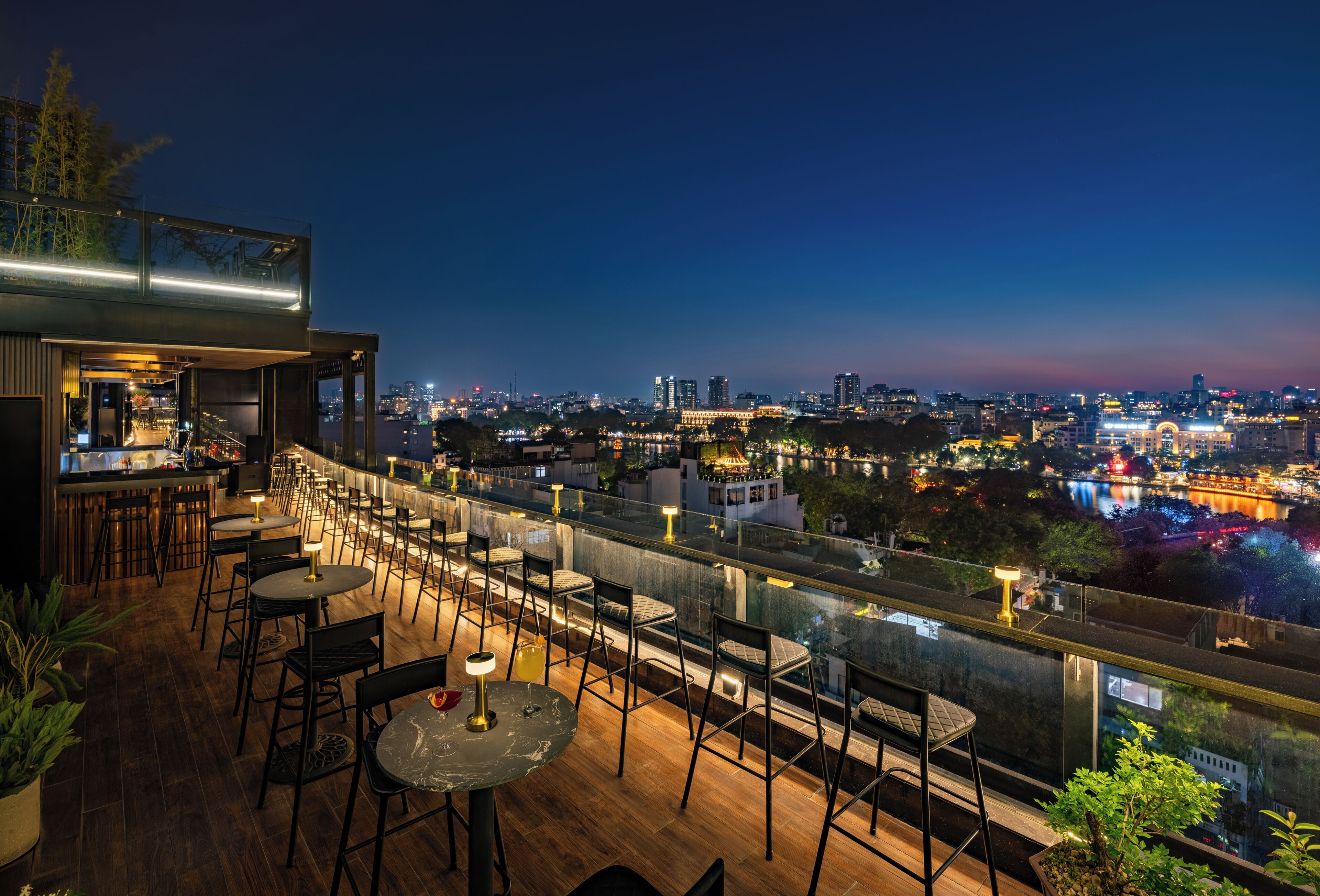 A rooftop bar with tables and chairs, overlooking a cityscape at dusk.