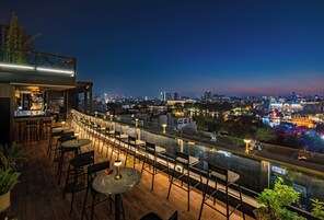 A rooftop bar with tables and chairs, overlooking a cityscape at dusk.