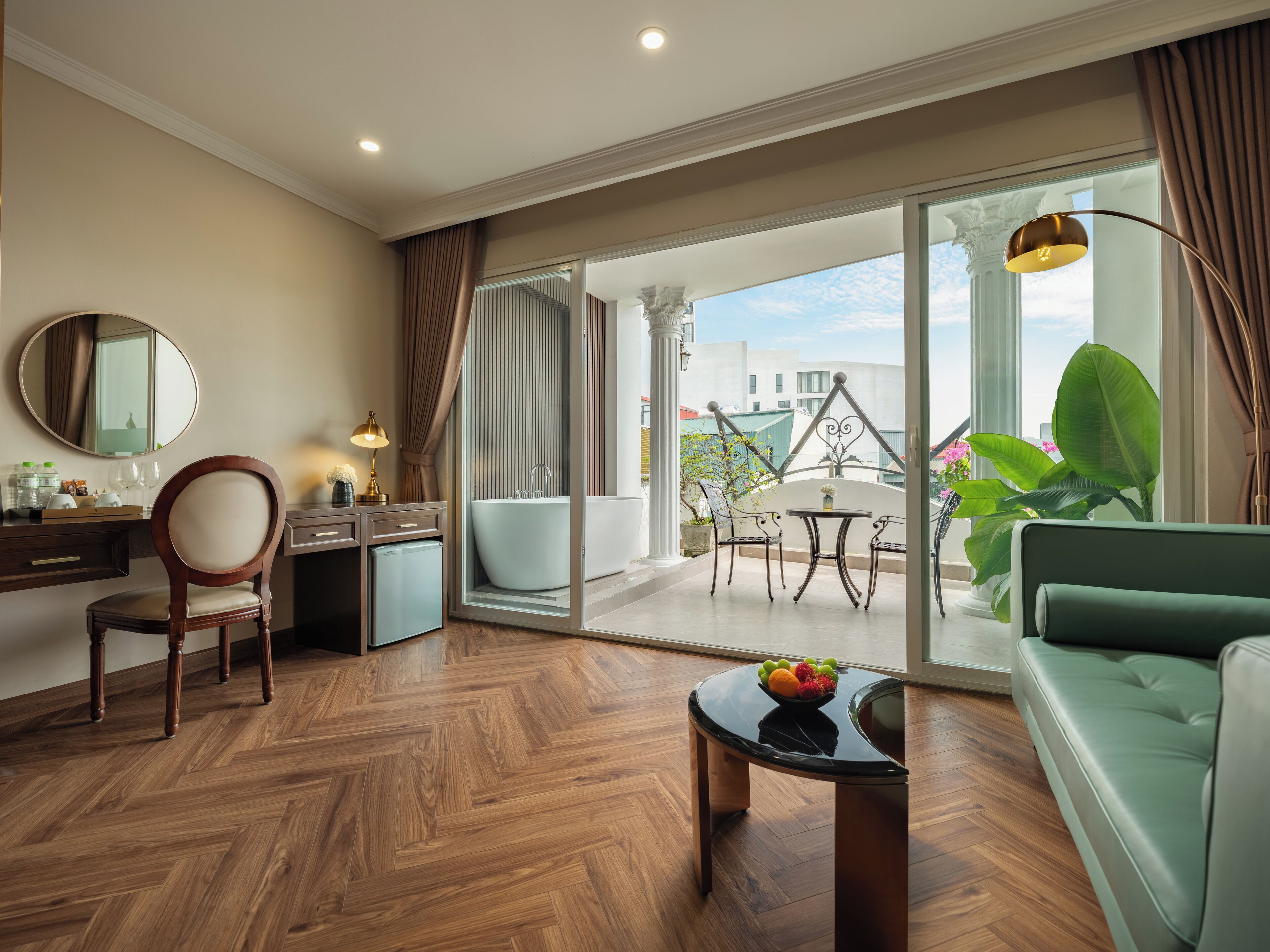 A modern hotel room with a wooden herringbone floor, a green sofa, a dining table with fruit, a desk with a mirror, and a view of the city through large windows.