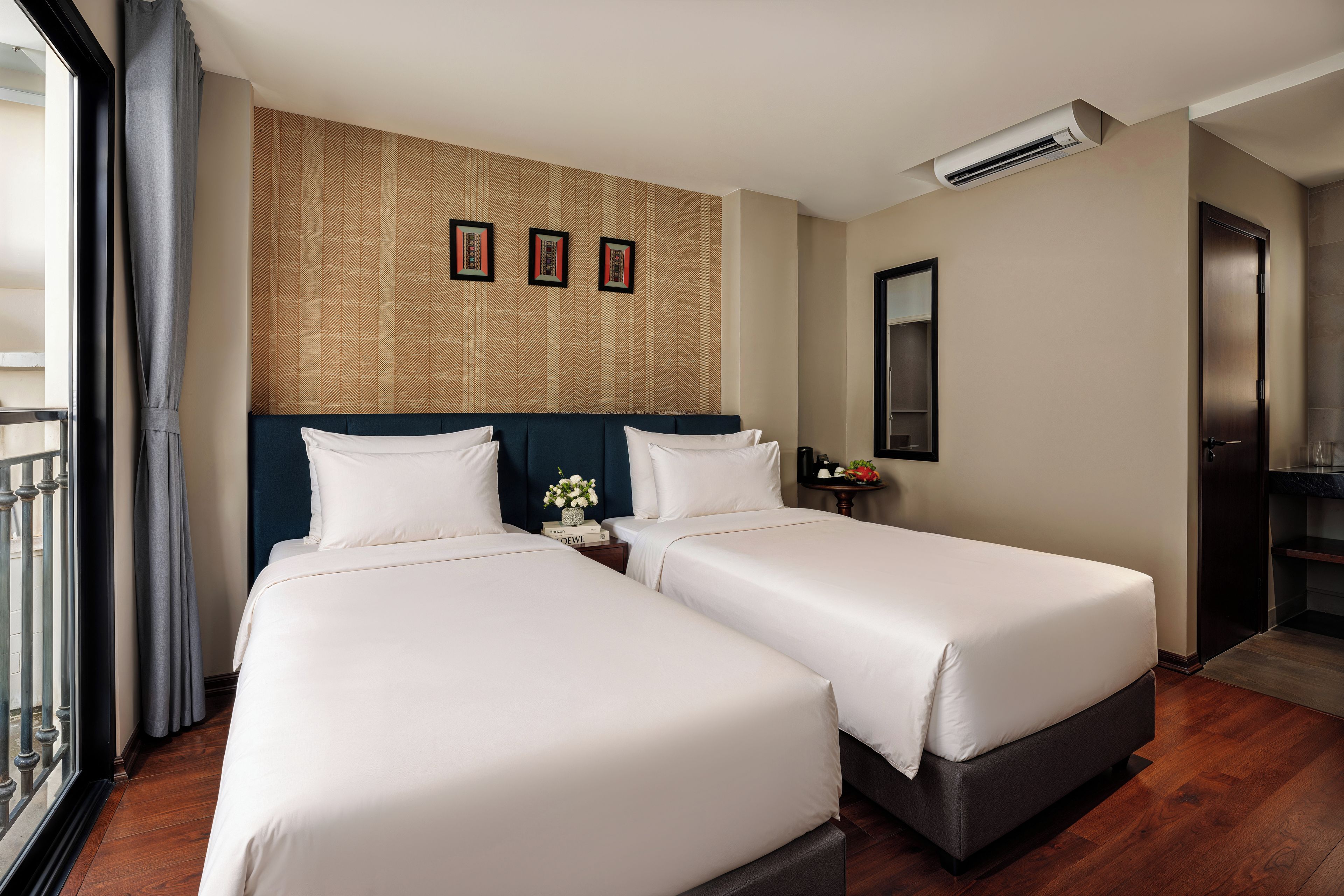 A hotel room with two beds, a patterned wall, a small table with a vase, and a door.