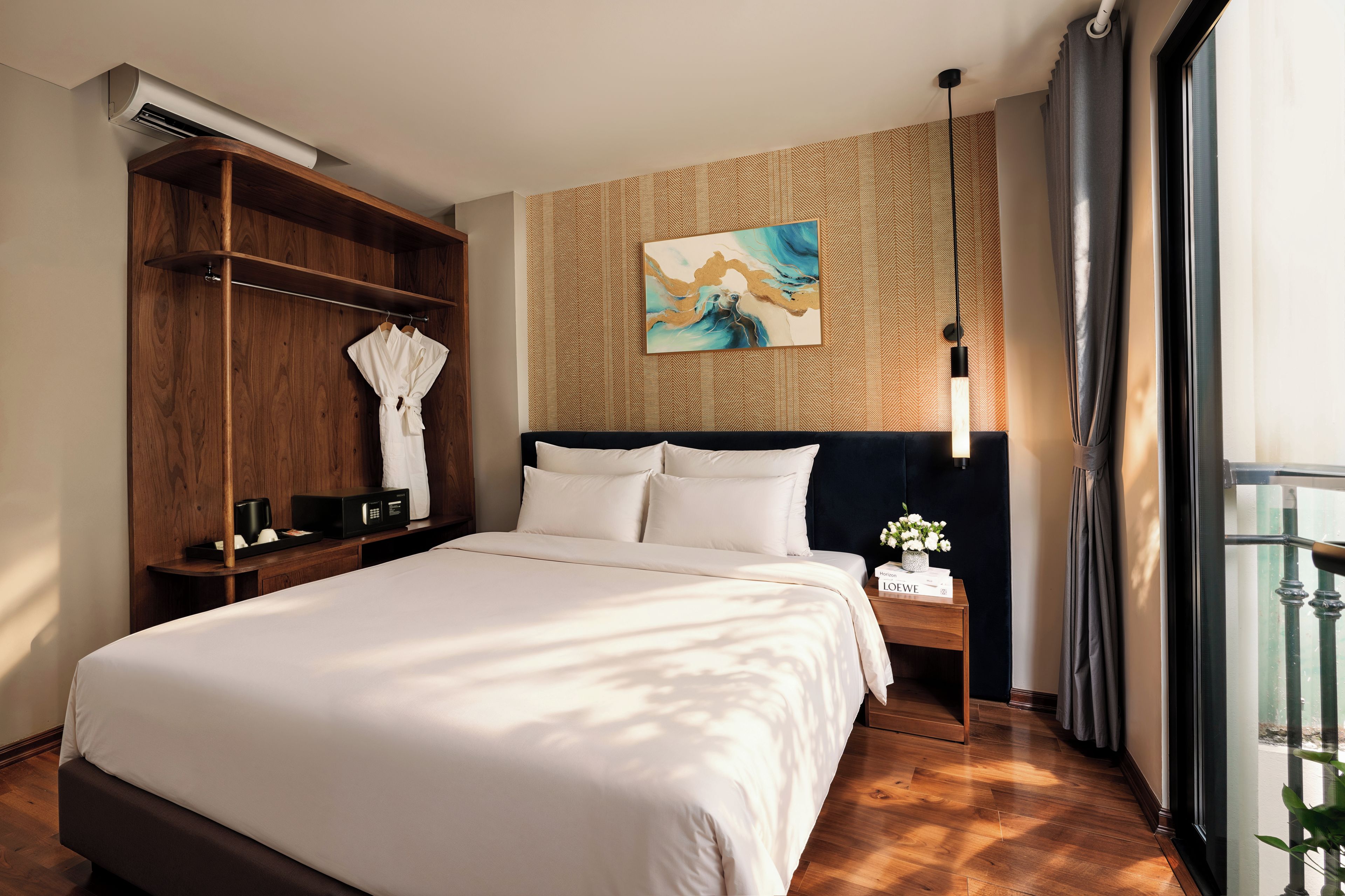 A hotel room with a large bed, a wooden wardrobe, a painting on the wall, and a nightstand with a lamp.