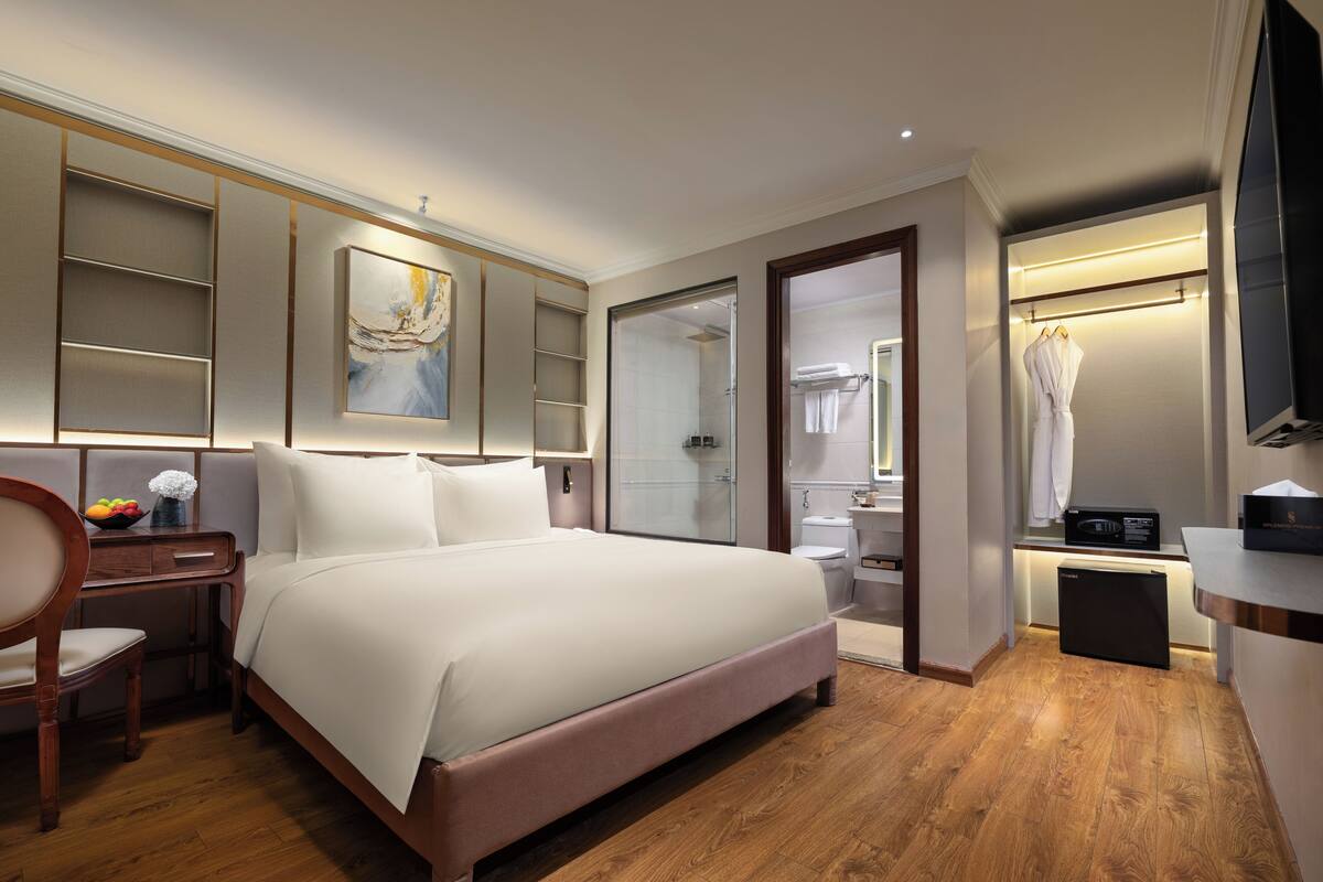 A modern hotel room with a large bed, a bedside table with a fruit bowl, a mirror, a shower area, and a TV.