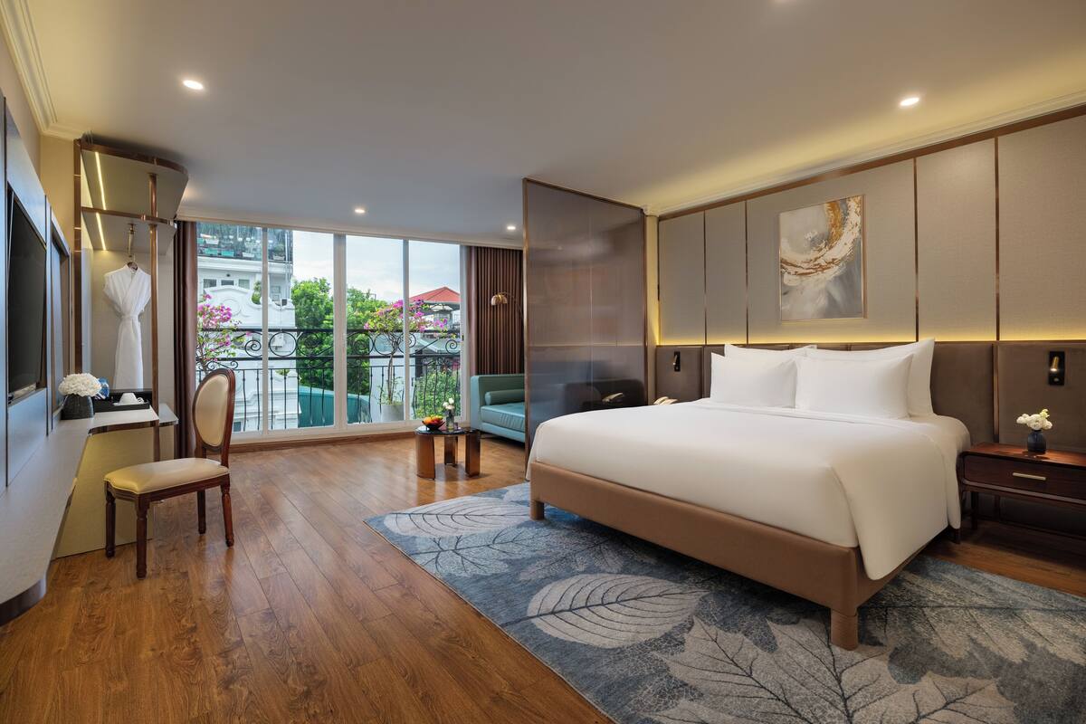 A spacious bedroom with a large bed, a sitting area with a chair, a desk, and a view of the outdoors.