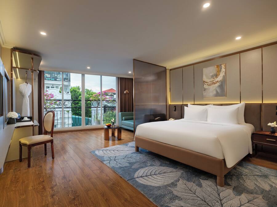 A spacious bedroom with a large bed, a sitting area with a chair, a desk, and a view of the outdoors.