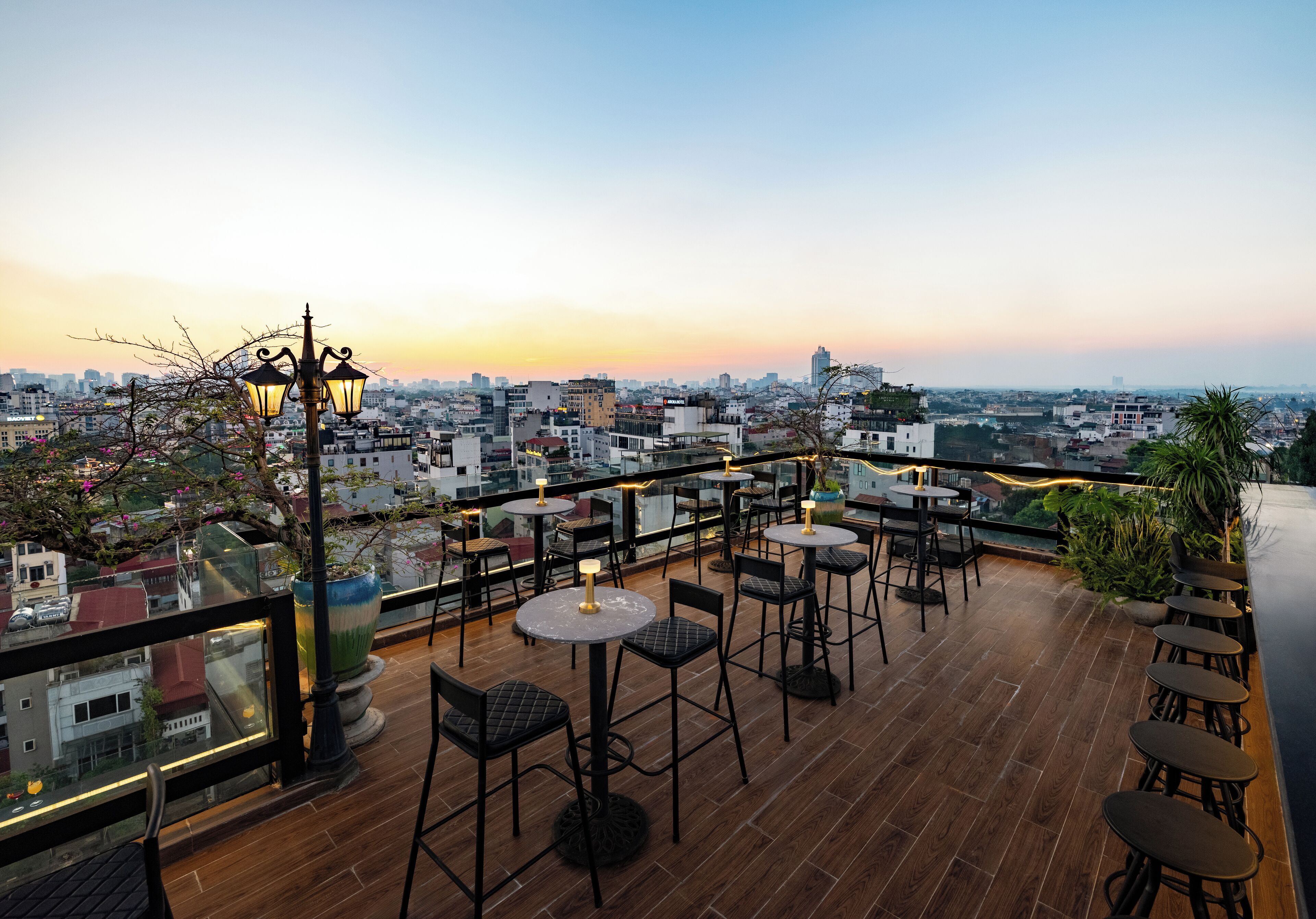 A rooftop terrace with tables and chairs, a street view, and a sunset in the background.