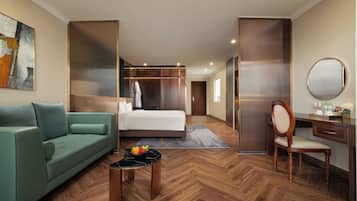 Una habitación de hotel con una cama, un sofá verde, un escritorio con una silla, un espejo redondo y un frutero sobre una mesita.