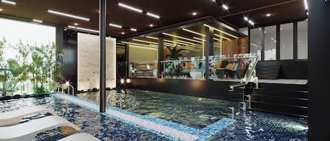 Una piscina interior moderna con agua cristalina, barandilla de vidrio y vista a un área de restaurante.