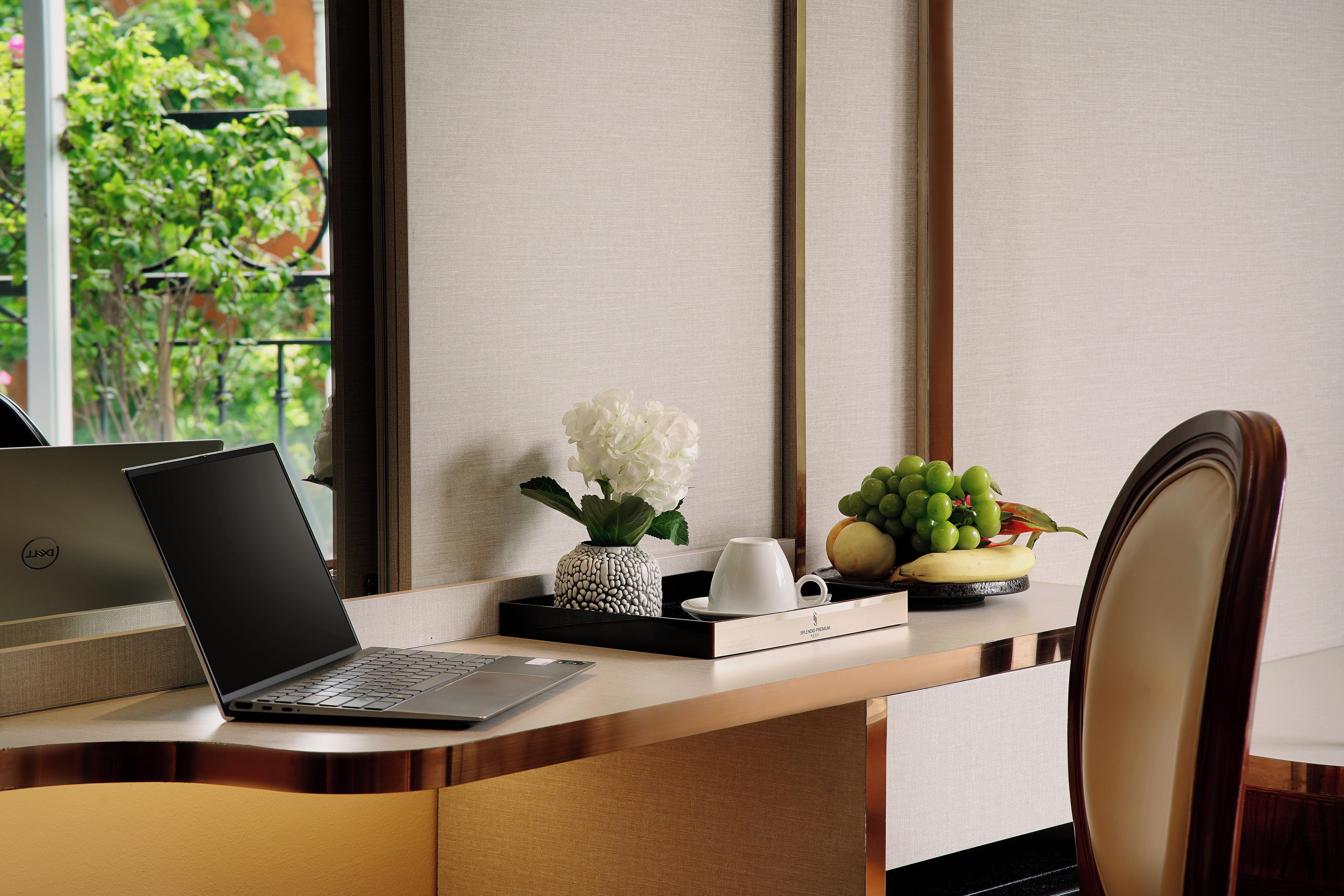 A laptop on a desk with a vase of flowers, a cup, and a fruit bowl.