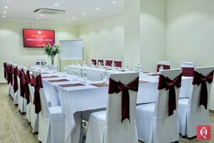 Un salón de conferencias con mesas y sillas blancas, cintas rojas y una pantalla mostrando un mensaje de bienvenida.
