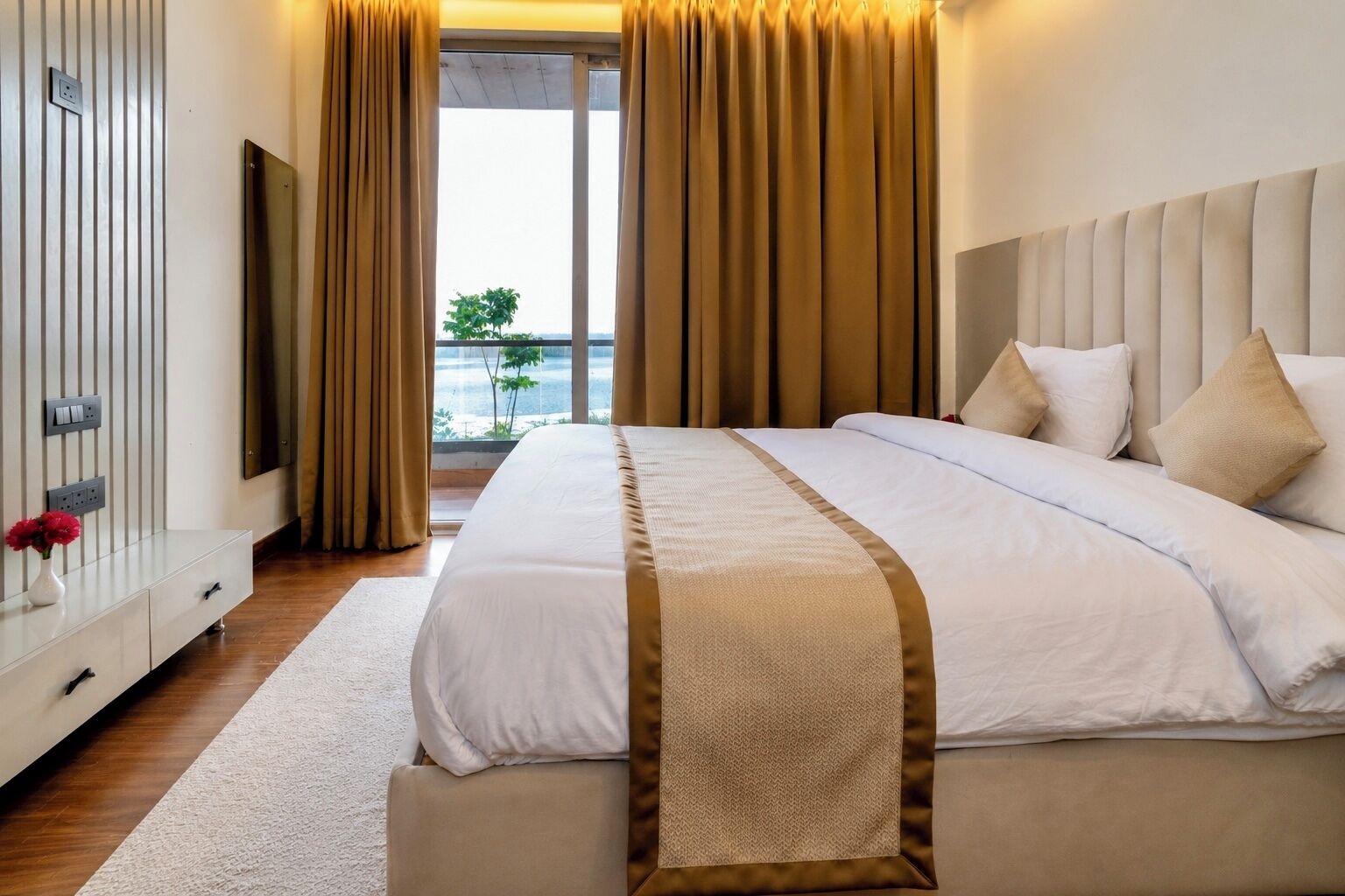 Una habitación de hotel con cama, mesita de noche con un jarrón de flores, ventana grande con cortinas y vistas al exterior.