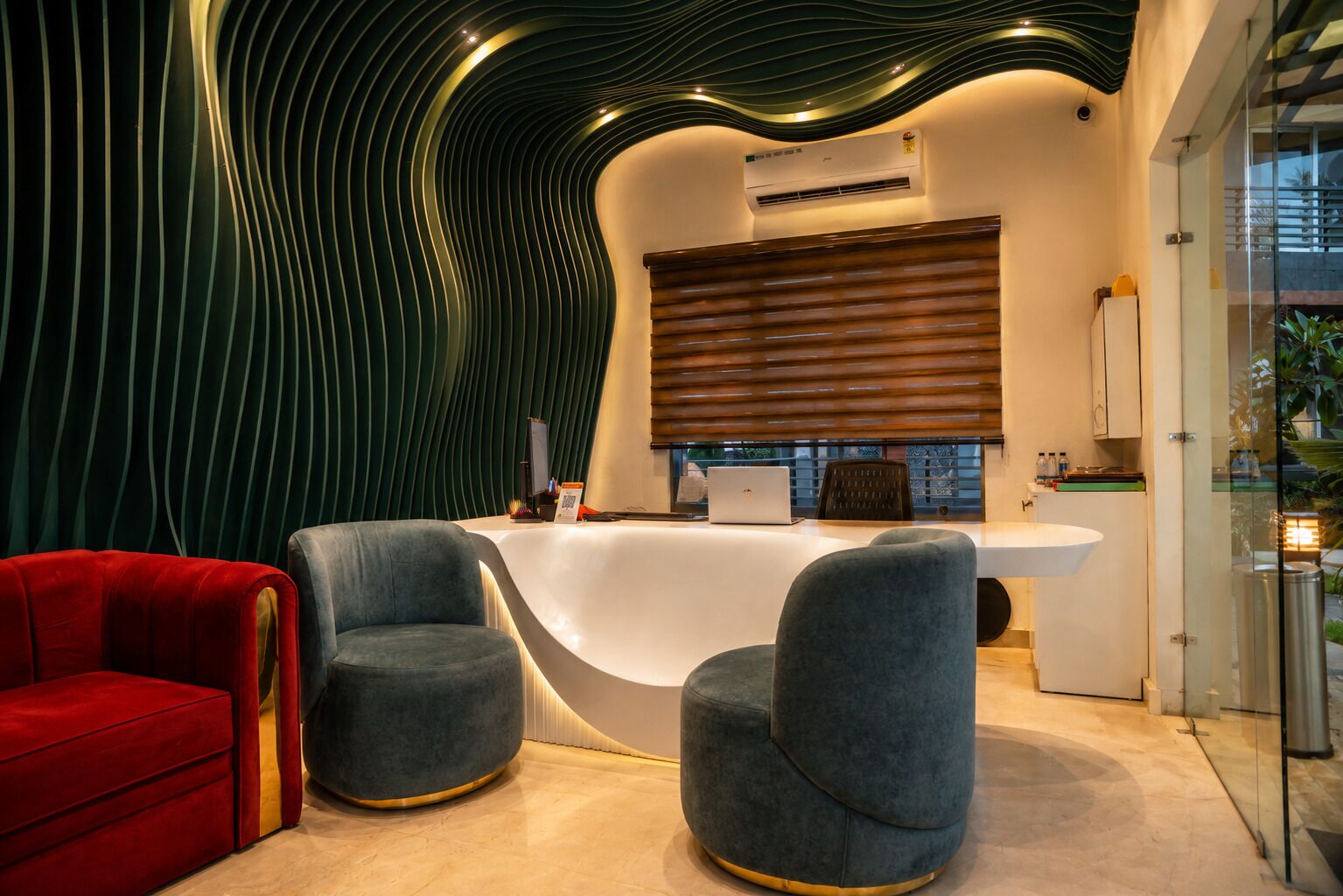 A modern interior with a curved reception desk, two grey armchairs, a red sofa, and a unique wavy ceiling design.